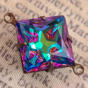 May include: A square, iridescent jewel with a bronze-colored metal setting. The jewel has a vibrant color scheme of purple, teal, and green. The setting includes loops on two sides. The jewel is likely a decorative element for jewelry or crafts.