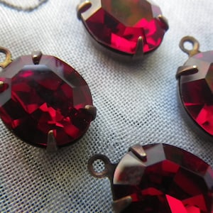 Puede incluir: Primer plano de varios colgantes de vidrio rojo oscuro facetados y de forma ovalada, con engastes y bucles de metal color bronce. Los colgantes están dispuestos sobre un fondo de tela azul claro texturizada. Estos colgantes son adecuados para la fabricación de joyas.