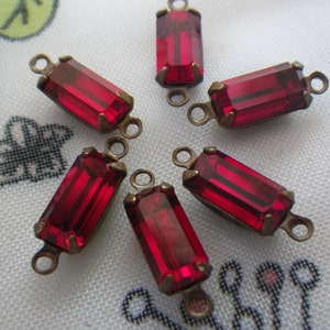 May include: Six rectangular red glass crystal connectors with brass-colored metal loops. The crystals have a faceted design, and the metal settings have small prongs. These components are suitable for jewelry making.