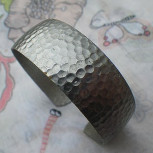 Könnte beinhalten: Ein silbernes, gehämmertes Armband aus Metall. Das Armband ist aus dickem Metall gefertigt und hat eine strukturierte Oberfläche.