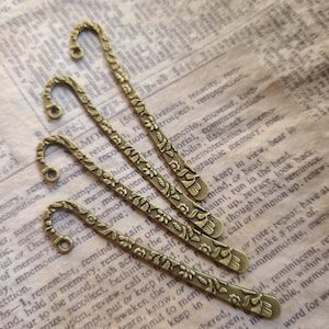 May include: Four antique brass-colored metal bookmarks with a floral design. Each bookmark has a curved top with a small hole and a decorative floral pattern along the length. The bookmarks are arranged on a vintage paper background.