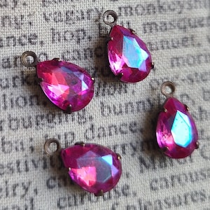 May include: Four pink iridescent teardrop-shaped glass charms with antique bronze metal loops. The charms are on a textured fabric background.