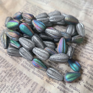 Peut inclure: Un ensemble de perles en forme de goutte. Les perles sont principalement grises avec des reflets irisés bleus, verts et violets. Les perles ont une surface texturée et striée. Les perles sont dispersées sur un fond de papier vintage.