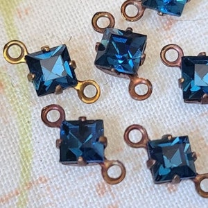 Könnte beinhalten: Sechs blaue quadratische Glasverbinder mit goldenen Metallschlaufen. Die Verbinder befinden sich auf einem weißen Stoffhintergrund.