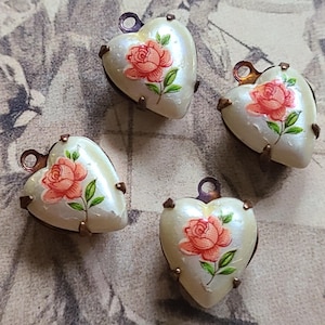 May include: Four heart-shaped pendants with a pearlescent finish. Each pendant features a painted pink rose with green leaves. The pendants have bronze-colored metal accents and a small loop at the top for hanging.