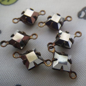 May include: Close-up of six faceted, square-shaped crystal connectors with bronze-colored metal settings and loops. The crystals reflect light, and the connectors are arranged on a patterned fabric.
