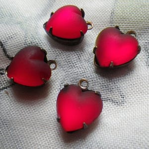 Puede incluir: Cuatro dijes rojos en forma de corazón con un acabado esmerilado y un engaste de metal color bronce. Cada dije tiene un pequeño lazo en la parte superior para sujetarlo a la joyería. Los dijes están dispuestos sobre una superficie blanca texturizada.