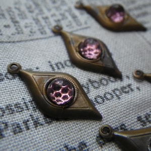 Può includere: Sei ciondoli in ottone antico a forma di lacrima, ognuno con un piccolo cabochon rotondo in vetro viola al centro. I ciondoli hanno un anello nella parte superiore per essere attaccati a una collana o un braccialetto.