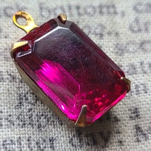 May include: A rectangular pendant with a faceted, deep pink gemstone. The gem is set in a gold-toned metal frame with a small loop at the top for a chain. The pendant has a vintage aesthetic.