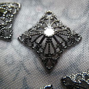 May include: A close-up of a silver filigree embellishment with a square shape and intricate details. The embellishment has a delicate, lacy design and a shiny finish.