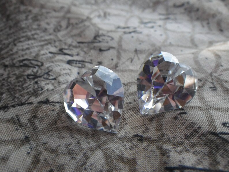 May include: Two clear crystal heart-shaped charms, with a faceted design, are displayed on a textured fabric background.