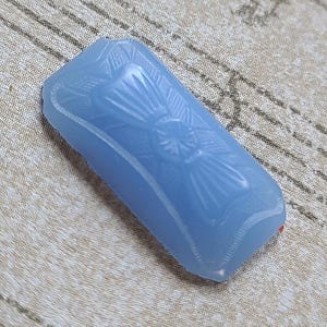 May include: A light blue, rectangular pendant with an Art Deco design. The pendant features a central floral motif with radiating lines and a curved, beveled edge. The surface is smooth and translucent, with a slight sheen.