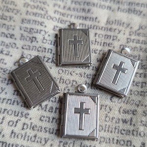 May include: Four silver-toned metal book charms with a cross design. Each charm is hinged and opens to reveal a small space inside.
