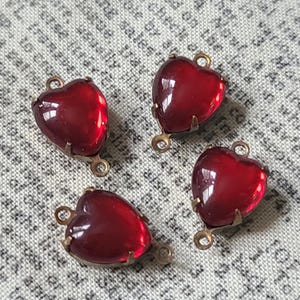 Puede incluir: Cuatro dijes de vidrio rojo en forma de corazón con engastes de metal color latón. Cada dije tiene un lazo en la parte superior y un pequeño lazo en la parte inferior para sujetarlo a joyas u otras manualidades. El vidrio es de un rojo profundo y translúcido.