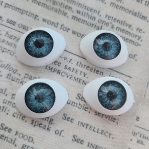 Pode incluir: Quatro olhos de boneca com íris azul e pupila preta, emoldurados em uma moldura oval branca. Os olhos têm detalhes intrincados e estão dispostos em um fundo de papel vintage.