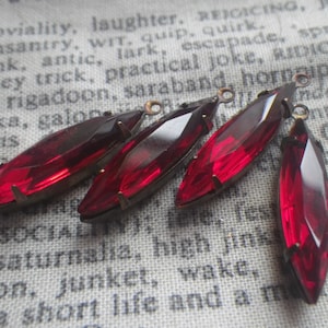 May include: Five red glass teardrop-shaped pendants with metal loops. The pendants are on a white fabric background.