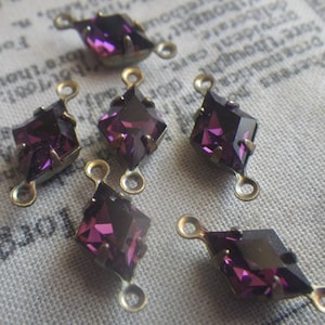 May include: Six faceted, diamond-shaped purple crystal charms with gold-colored metal loops. The crystals have a deep purple hue with a reflective surface. These charms are ideal for jewelry making or other craft projects.