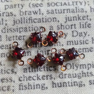 May include: Six small, dark red gemstone connectors with copper-colored loops. The gemstones are square-cut and faceted, catching the light. The connectors are arranged on a textured, off-white fabric with faded text.