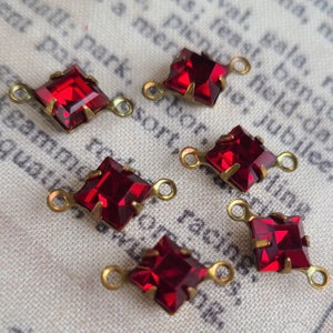 May include: Close-up of five red square-cut crystal charms with brass-colored metal settings and loops. The crystals are faceted, catching the light. The charms are on a patterned fabric background.