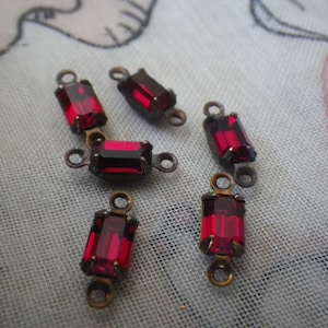 May include: Six red glass rectangular-shaped beads with gold-toned metal loops on each end. The beads are on a white fabric surface.