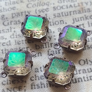 May include: Four square-cut iridescent glass beads with a metallic setting. Each bead has a central green hue with a faceted design, surrounded by a shimmering, multi-colored effect. The beads have small loops for attachment.