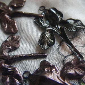 May include: Close-up of several dark bronze-colored iris flower charms. The charms have detailed petal designs and small loops for attaching to jewelry or other crafts. The background is a soft, patterned fabric.
