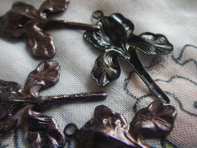 May include: Close-up of several dark bronze-colored iris flower charms. The charms have detailed petal designs and small loops for attaching to jewelry or other crafts. The background is a soft, patterned fabric.