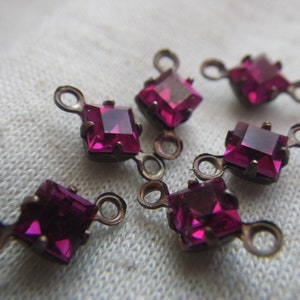 May include: Six pink square-shaped glass rhinestones with metal loops on each side. The rhinestones are on a white fabric surface.