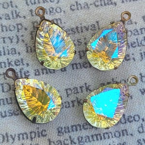 May include: Four teardrop-shaped pendants featuring iridescent, faceted glass stones set in antique bronze-toned metal frames. Each pendant has a small circular loop at the top for attachment, displaying a vibrant yellow and blue color shift.