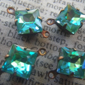 May include: Four square-cut, iridescent teal glass jewels with copper-colored metal settings and loops. The jewels have a faceted surface that reflects light, creating a shimmering effect. Ideal for jewelry making or embellishments.