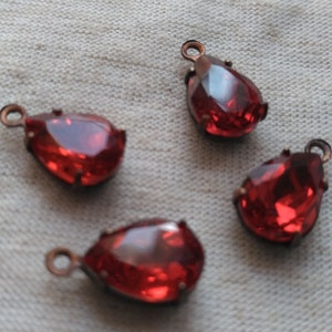 May include: Four red teardrop-shaped glass beads with copper-colored metal loops. The beads are on a white fabric background.