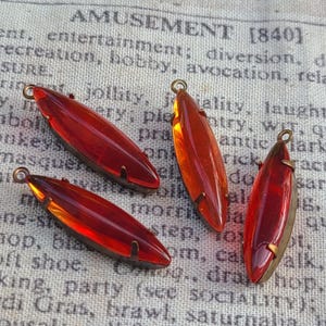 May include: Four elongated, teardrop-shaped pendants with a rich, reddish-orange hue. Each pendant has a metal frame and a small loop at the top for hanging. The pendants are displayed on a vintage fabric background.