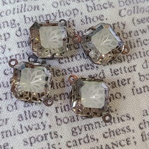 May include: Four square-cut, clear glass beads with etched floral designs. Each bead has a metal filigree frame and small loops for stringing. The beads are a light gray color and are displayed on a vintage fabric background.