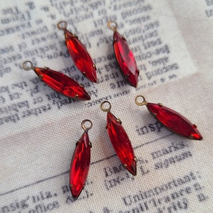 May include: Six red, marquise-shaped crystal pendants with brass-colored loops. The crystals are faceted and set in brass-colored metal. The pendants are arranged on a vintage fabric background with printed text.