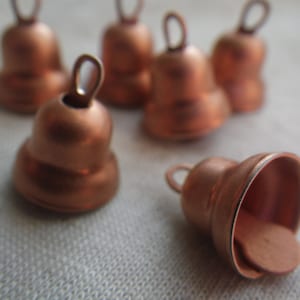 May include: Six small copper bells with a loop at the top. The bells are arranged in a semi-circle on a white surface.