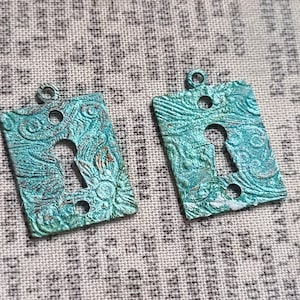 Może przedstawiać: Dwa prostokątne, turkusowe zawieszki w kształcie dziurki od klucza z teksturowanym, ozdobnym wzorem. Każda zawieszka ma wycięcie na dziurkę od klucza i małą pętlę u góry do zawieszenia. Zawieszki mają postarzany wygląd.