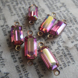 May include: Six iridescent pink glass rectangular beads with gold metal loops on each end. The beads have a faceted design and are arranged on a fabric surface.