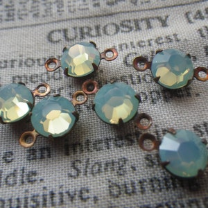 May include: Close-up of several opalescent blue-green faceted gemstone connectors with copper-colored metal loops. The gemstones have a yellow center and are set in a copper-colored metal. The background is a vintage newspaper print.