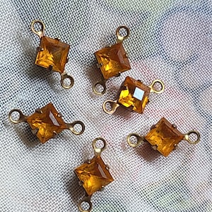 Könnte beinhalten: Sechs quadratische, bernsteinfarbene Glasanhänger im Vintage-Stil mit goldfarbenen Metallfassungen. Jeder Anhänger hat oben und unten eine Schlaufe zum Befestigen an Schmuck. Die Anhänger sind vor einem hellen, strukturierten Hintergrund platziert.