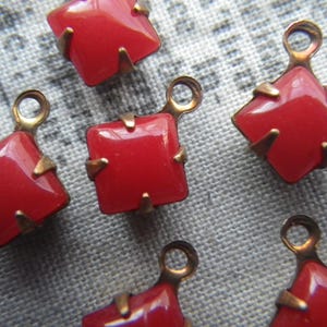 May include: Six red square glass rhinestones with gold metal settings. Each rhinestone has a small loop on the top for attaching to a project.