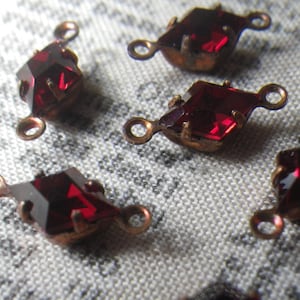 May include: Close-up of several faceted, dark red gemstone charms with copper-colored metal settings and loops for attaching to jewelry. The gems have a diamond shape.
