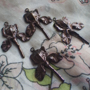 May include: Four bronze-colored iris flower charms with a loop at the top for jewelry making. The charms are detailed with petals and stems, and are arranged on a floral patterned fabric.