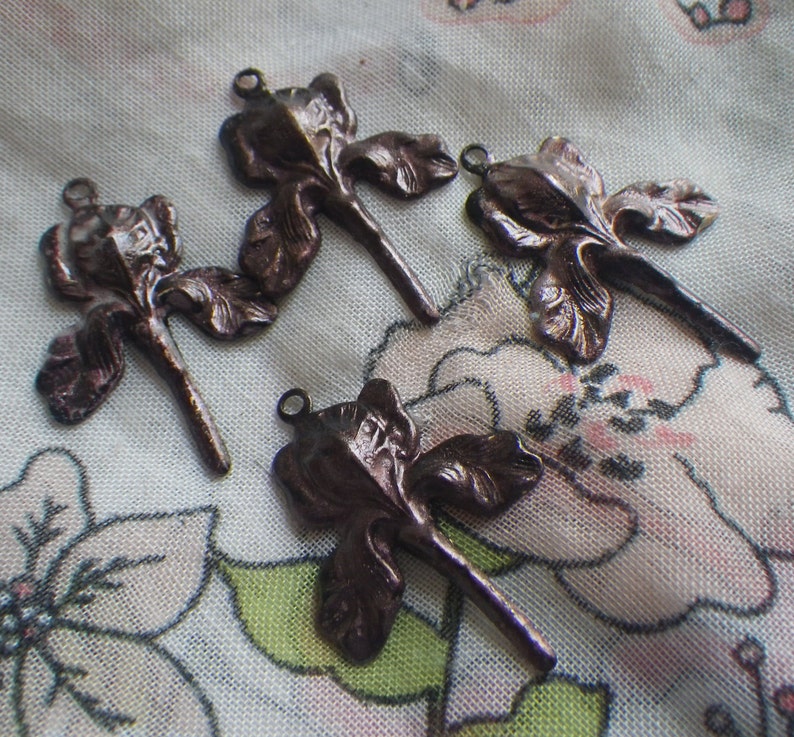 May include: Four bronze-colored iris flower charms with a loop at the top for jewelry making. The charms are detailed with petals and stems, and are arranged on a floral patterned fabric.