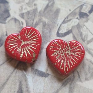 May include: Two red heart-shaped beads with gold-colored, sunburst-like designs. The beads are glossy and appear to be made of glass or a similar material. The background is a faded, vintage-style print.