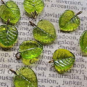 May include: A collection of small, translucent lime green glass leaf charms, each featuring a detailed vein pattern and a small bronze-toned metal jump ring attached to the top for hanging, arranged in a scattered pattern on a textured surface.