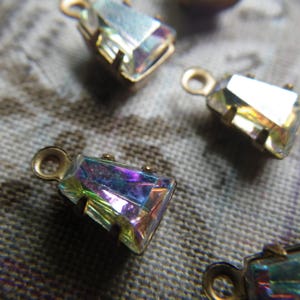 May include: Close-up of several iridescent rectangular crystal pendants with gold-coloured metal settings and small loops. The crystals reflect a spectrum of colours, including purple, blue, and yellow. The pendants are on a textured, neutral-toned surface.