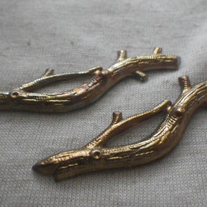 May include: Two gold-toned metal twig-shaped embellishments with a textured surface. The twigs are curved and have a branch-like design.