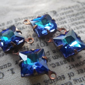 May include: Four blue square-shaped glass rhinestones with copper-coloured metal settings and loops. The rhinestones have a faceted design and a blue-green iridescent shimmer.