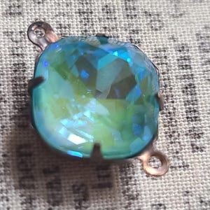 May include: A faceted, iridescent blue-green crystal pendant with a copper-colored metal setting. The crystal has a square shape with rounded edges and is designed with a loop at the top and bottom for attachment.