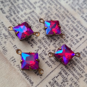May include: Four square, iridescent rhinestones with a vibrant pink and blue color scheme. Each rhinestone has a brass-colored metal setting with small loops on each corner, perfect for jewelry making or embellishments.